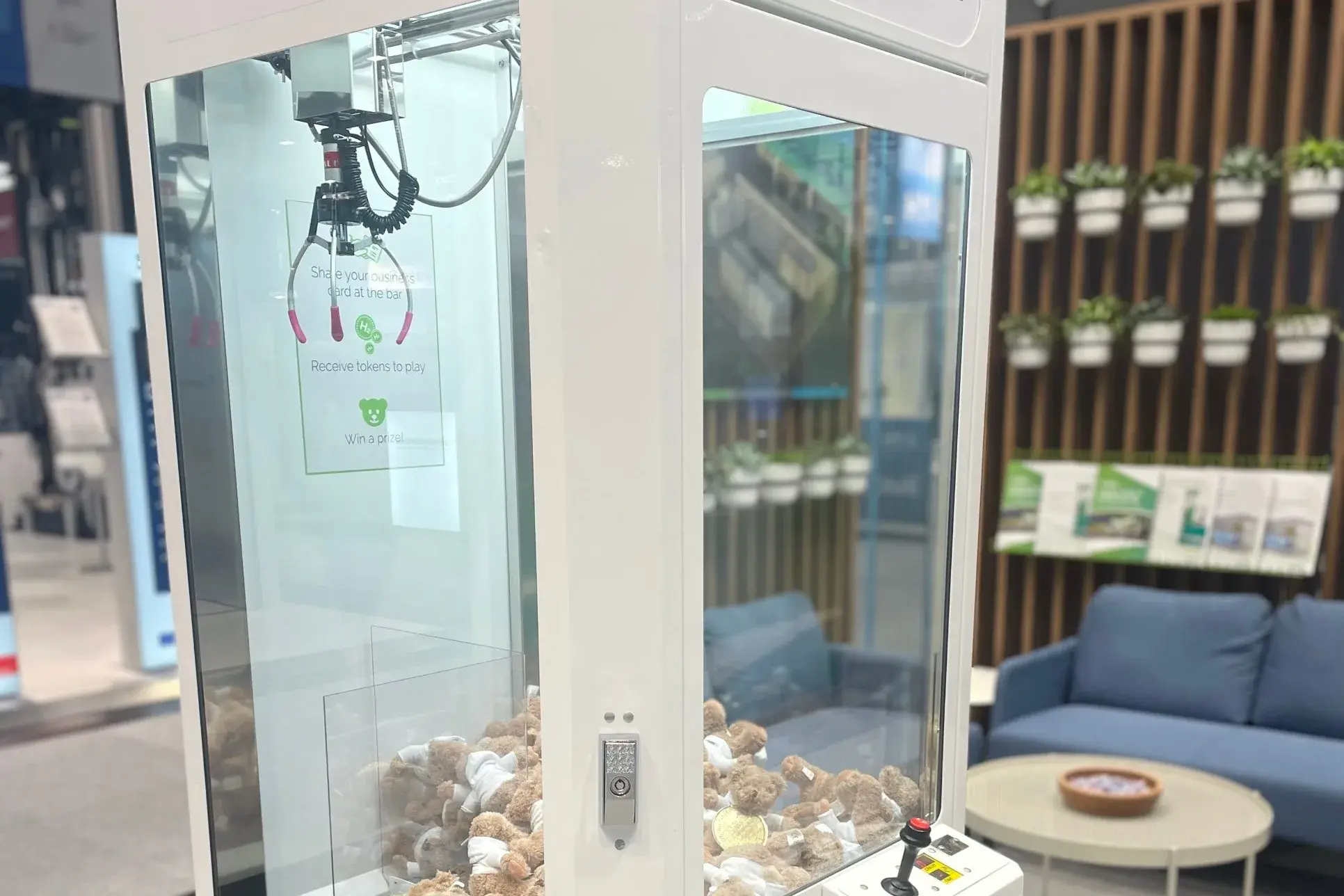 A white Gilbarco Veeder-Root H2 claw machine filled with teddy bears, with a card inviting visitors to swap a business card for tokens