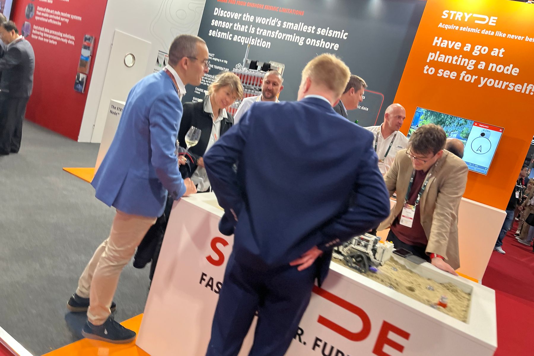 Delegates gathered around the sand bed under the orange Have a go at planting a node panel